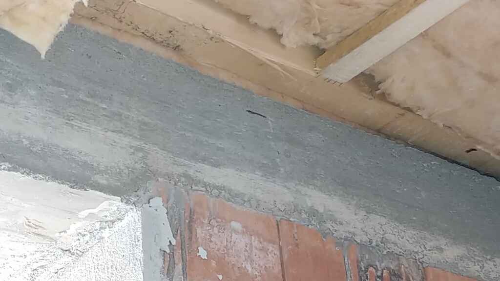 Innenansicht einer unfertigen Wand: grauer Putz, darunter Ziegel, Holzbalkendecke mit Dämmung.