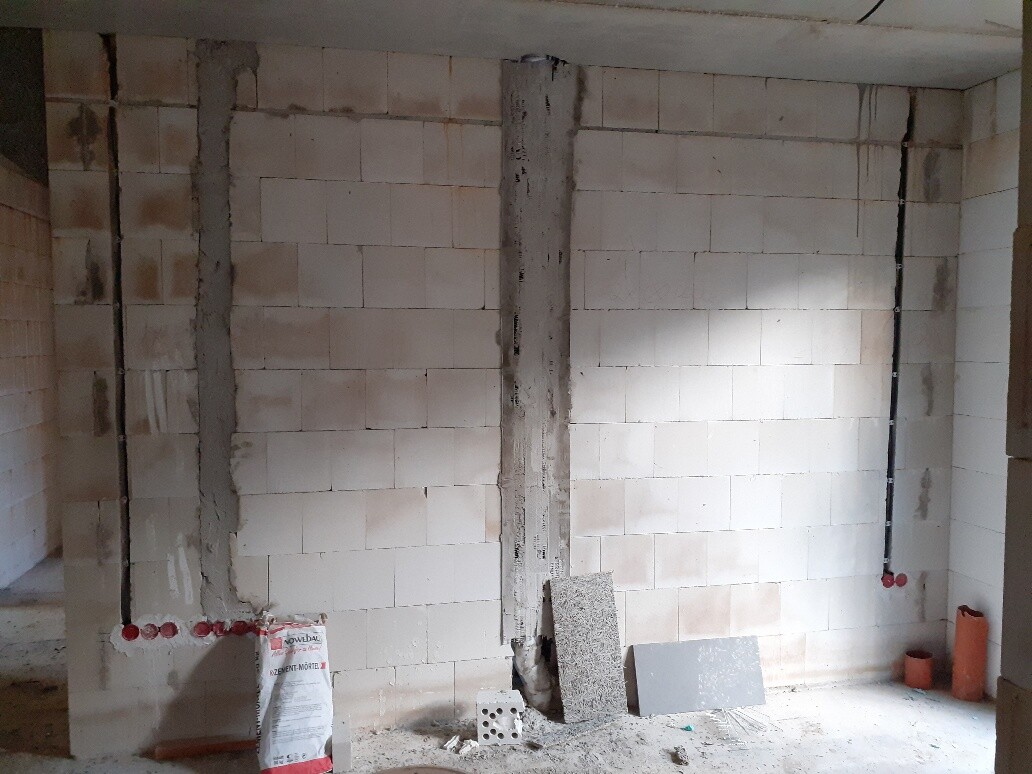 Unfertiger Innenraum mit Betonstütze, Wandblöcken, Rohrleitungen und Baumaterialien.