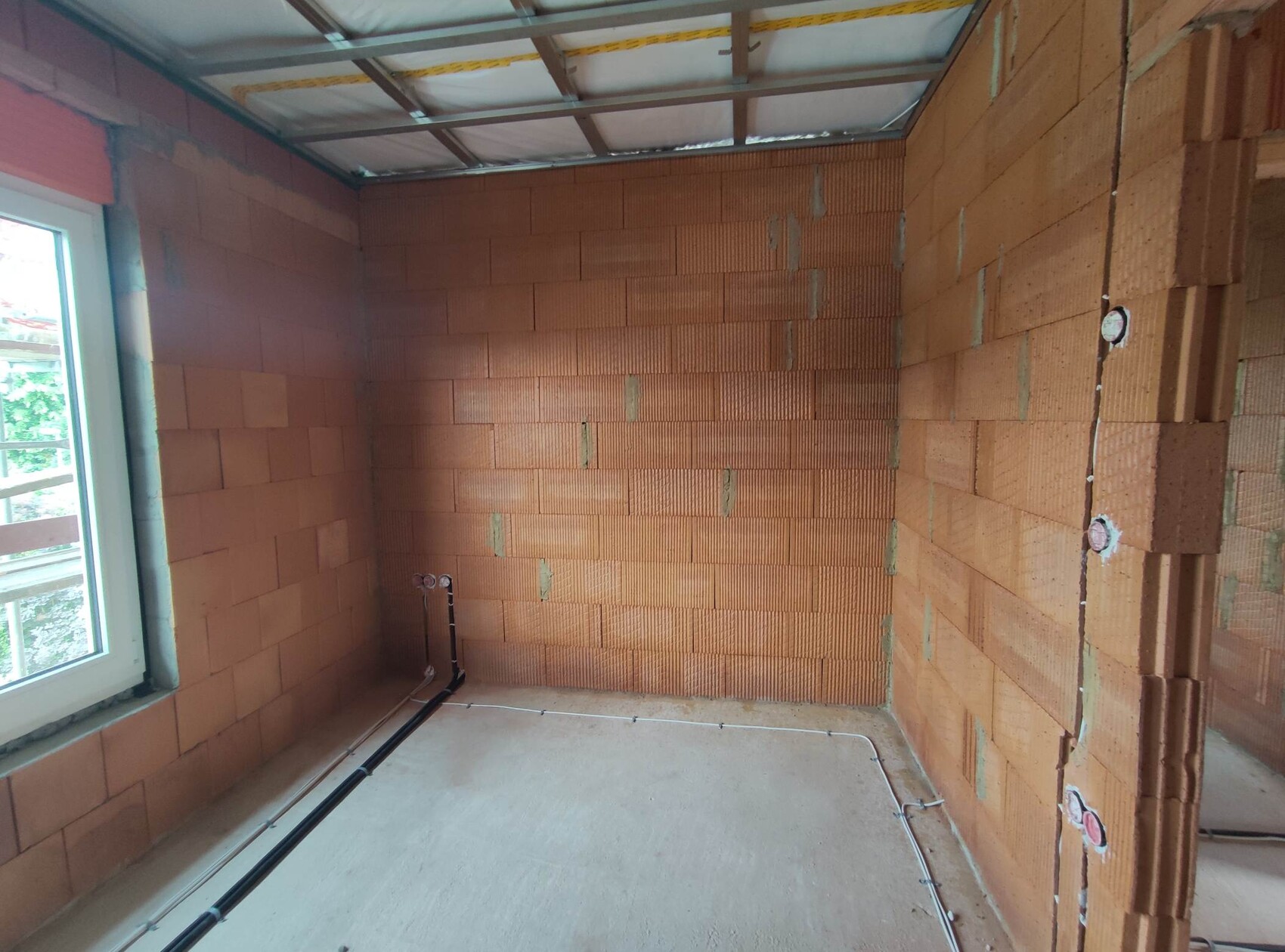 Rohbauzimmer mit roten Ziegelwänden, Fenster links, sichtbare Leitungen am Boden, offene Decke.
