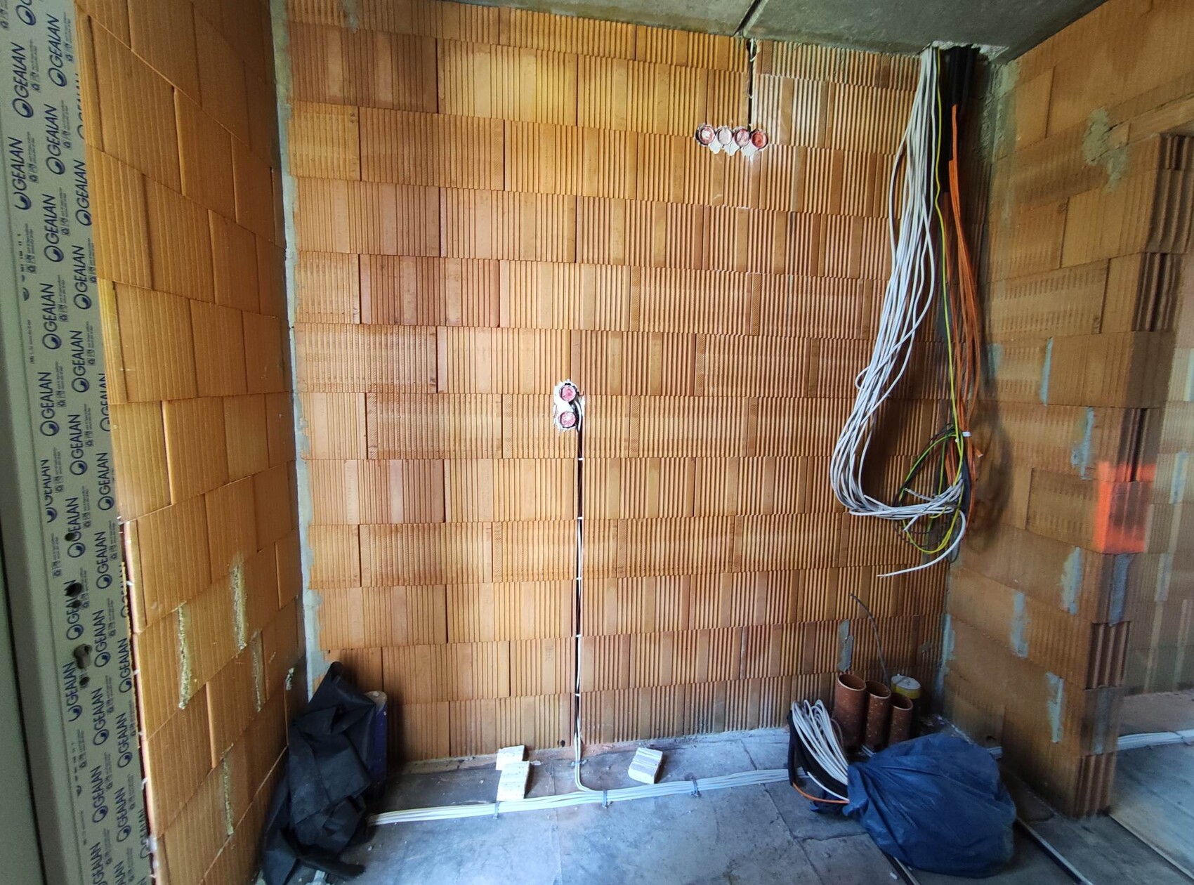 Innenraum einer Baustelle mit Ziegelwand, sichtbaren Kabelbündeln, Rohren und Bauteilen am Boden.