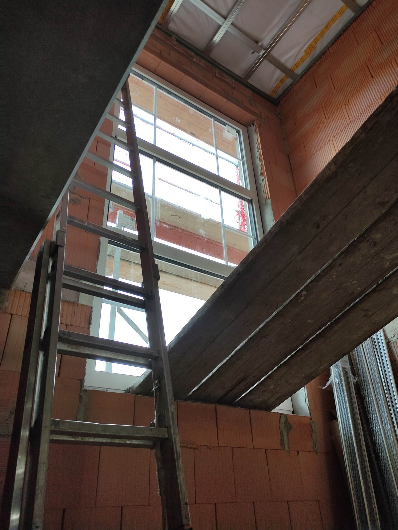 Innenansicht einer Baustelle: Metallleiter, Ziegelwand und große Fensteröffnung.