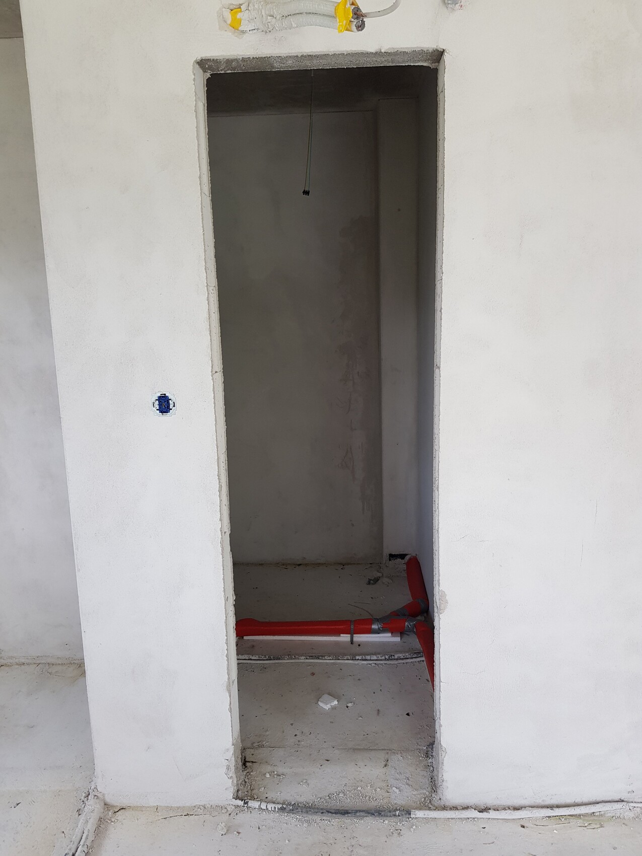 Unfertiger Innenraum: Türöffnung in weißer Wand, rotes Rohr am Boden, lose Kabel von der Decke.
