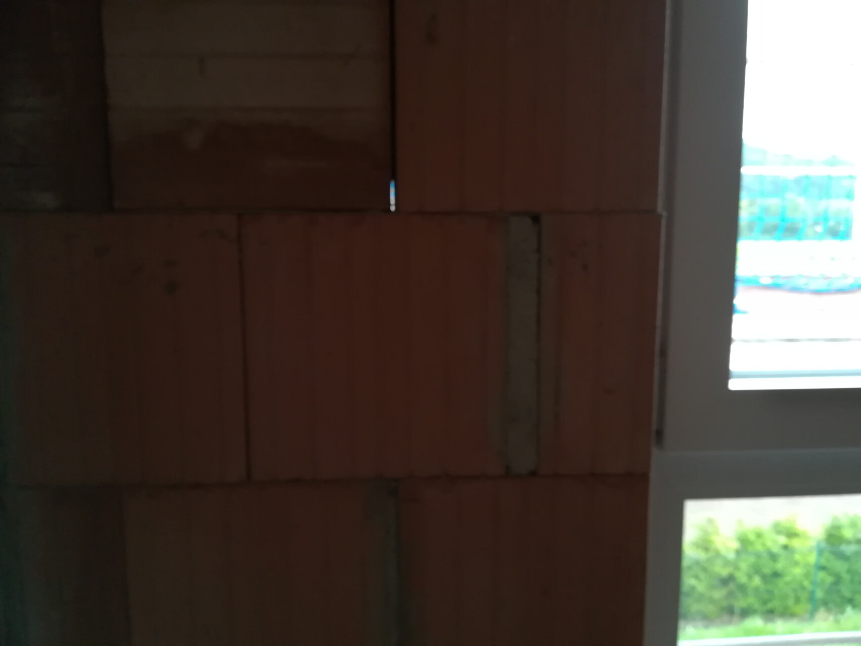 Dunkle Innenaufnahme einer roten Ziegelwand neben einem weißen Fenster; draußen grüne Hecke sichtbar.