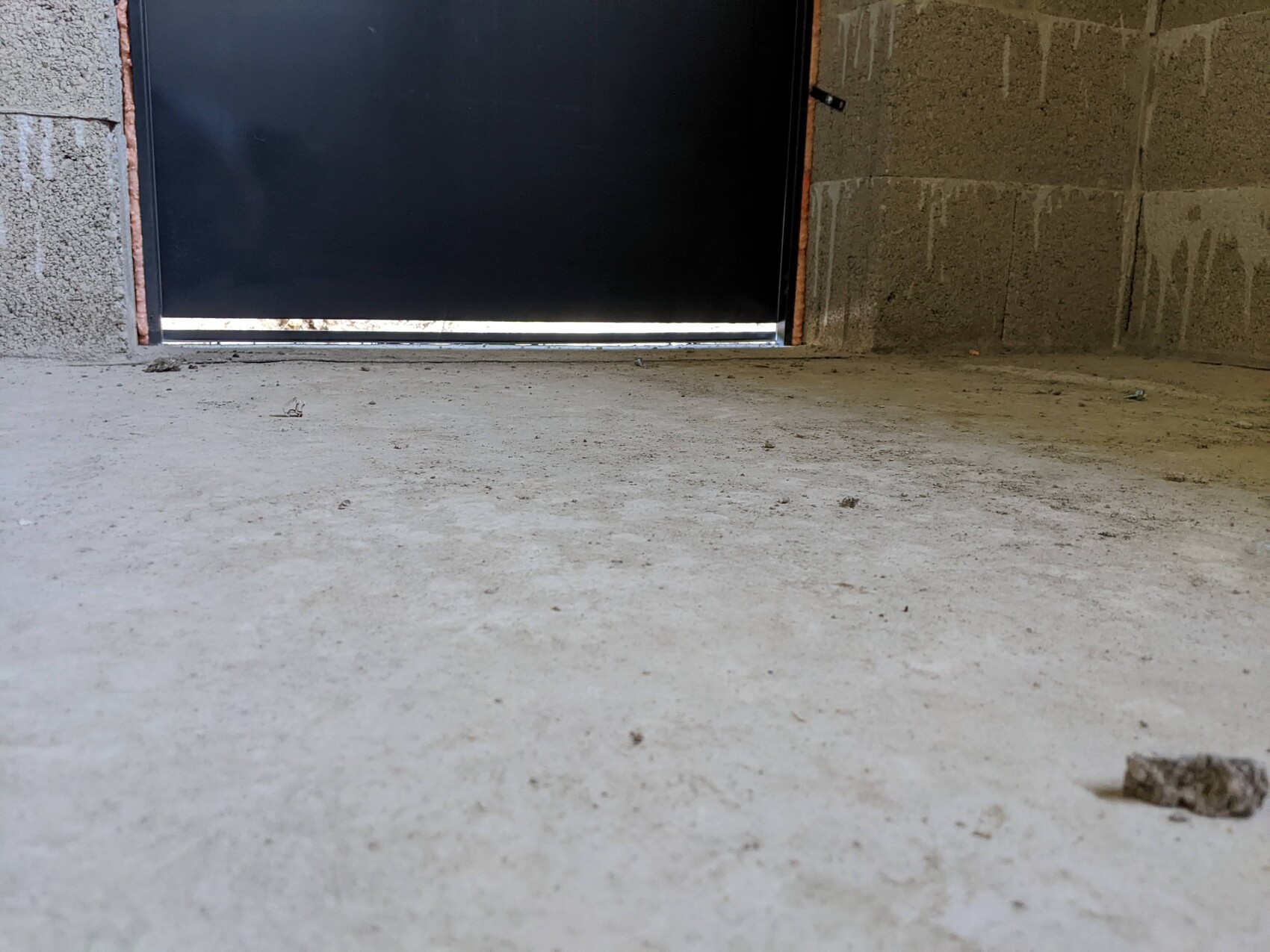 Bodennahes Bild einer dunklen Metalltür in einer rauen Betonwand, staubiger Boden, Lichtspalt unten