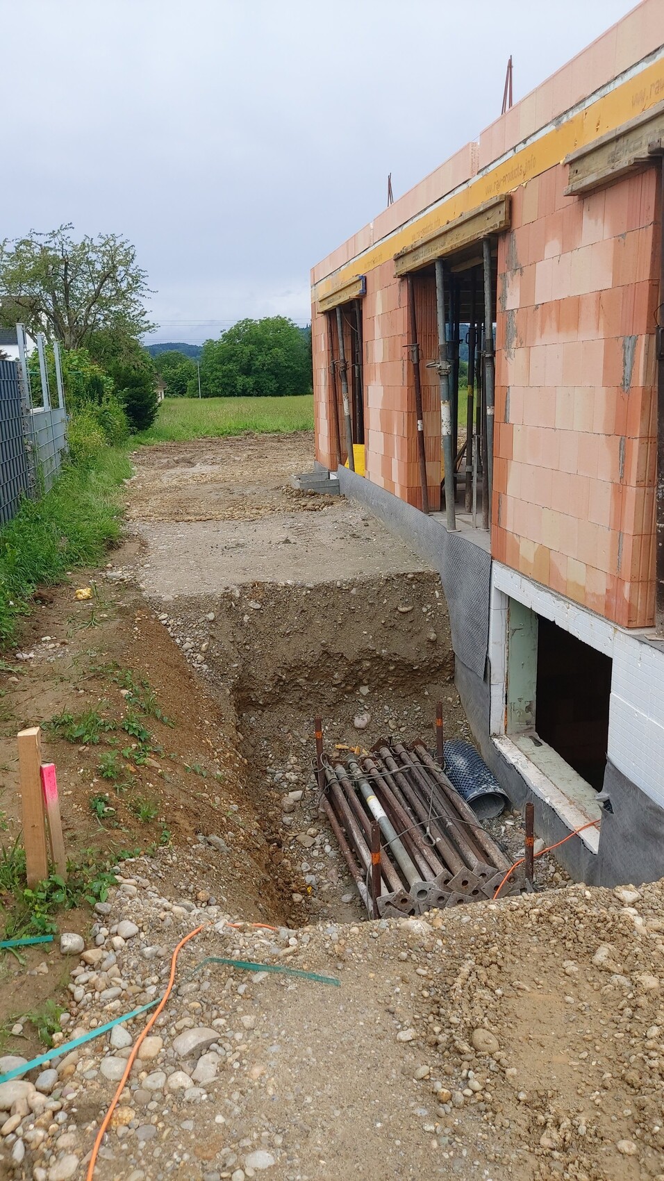 Baustelle: offenes Erdgrab neben rotem Ziegelhaus; Rohre und Bewehrung sichtbar.