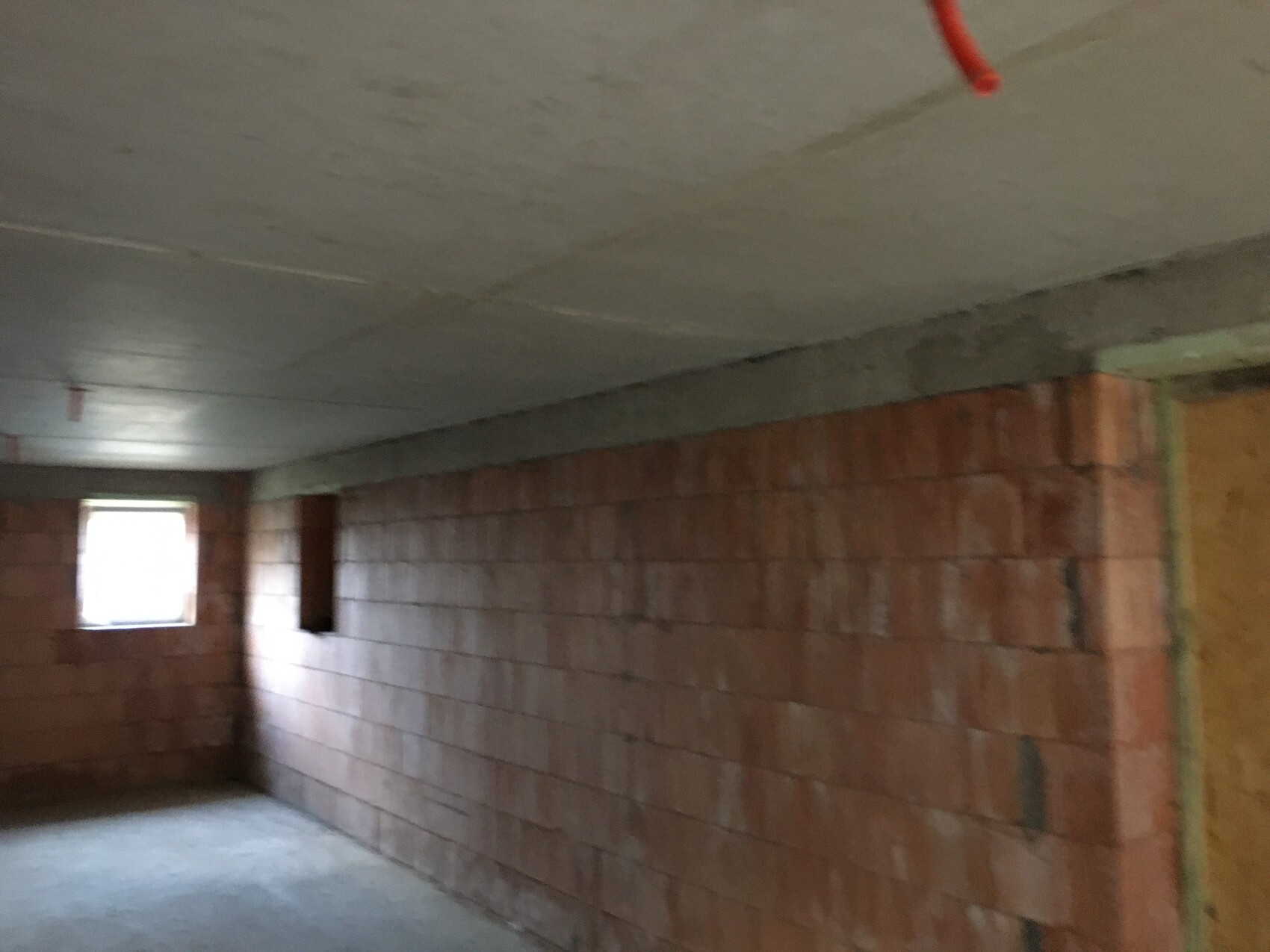 Unbearbeiteter Kellerraum mit Ziegelwänden, Betonboden/Decke, kleinem Fenster links, rotem Rohr.