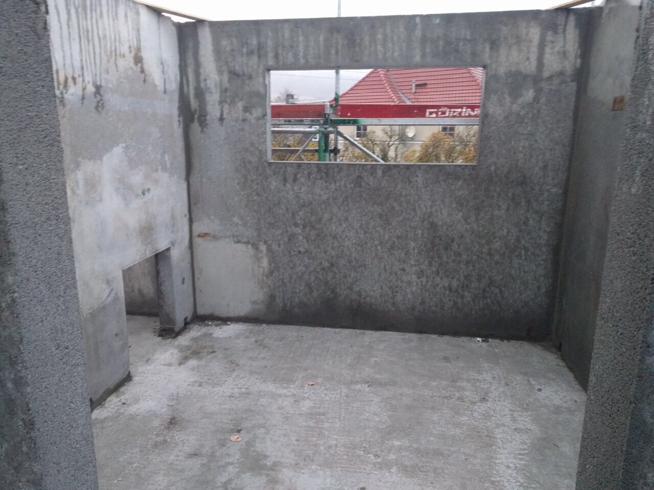 Innenansicht eines unfertigen Betonraums; Fensteröffnung zeigt Baugerüst und rotes Dach.
