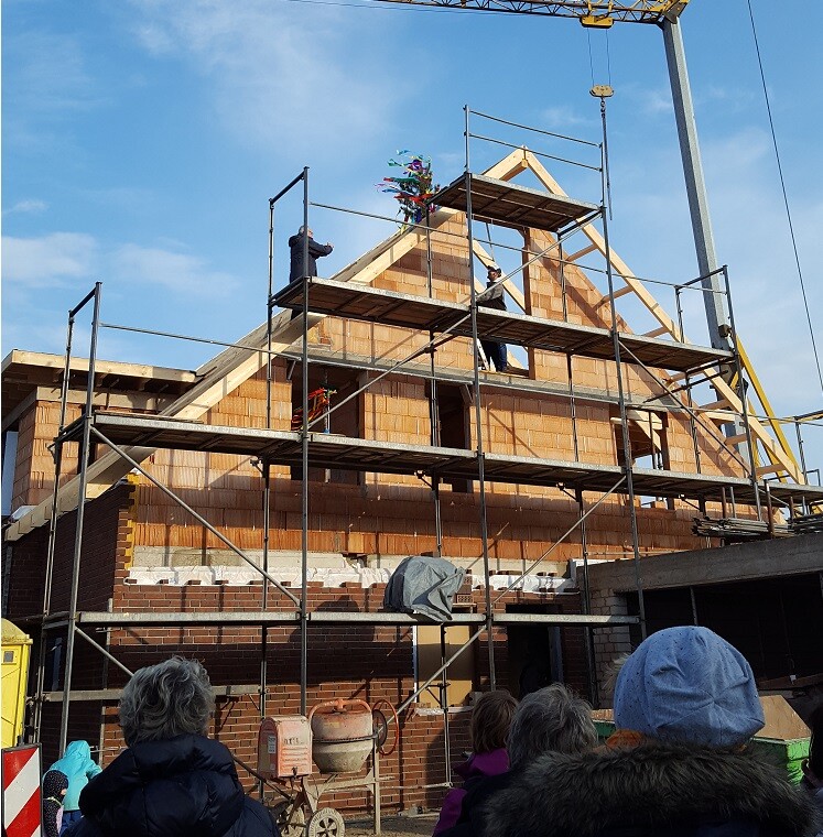 Holzhaus im Rohbau mit Gerüst; Arbeiter auf Plattform, bunter Dachfirst, Kran im Hintergrund.