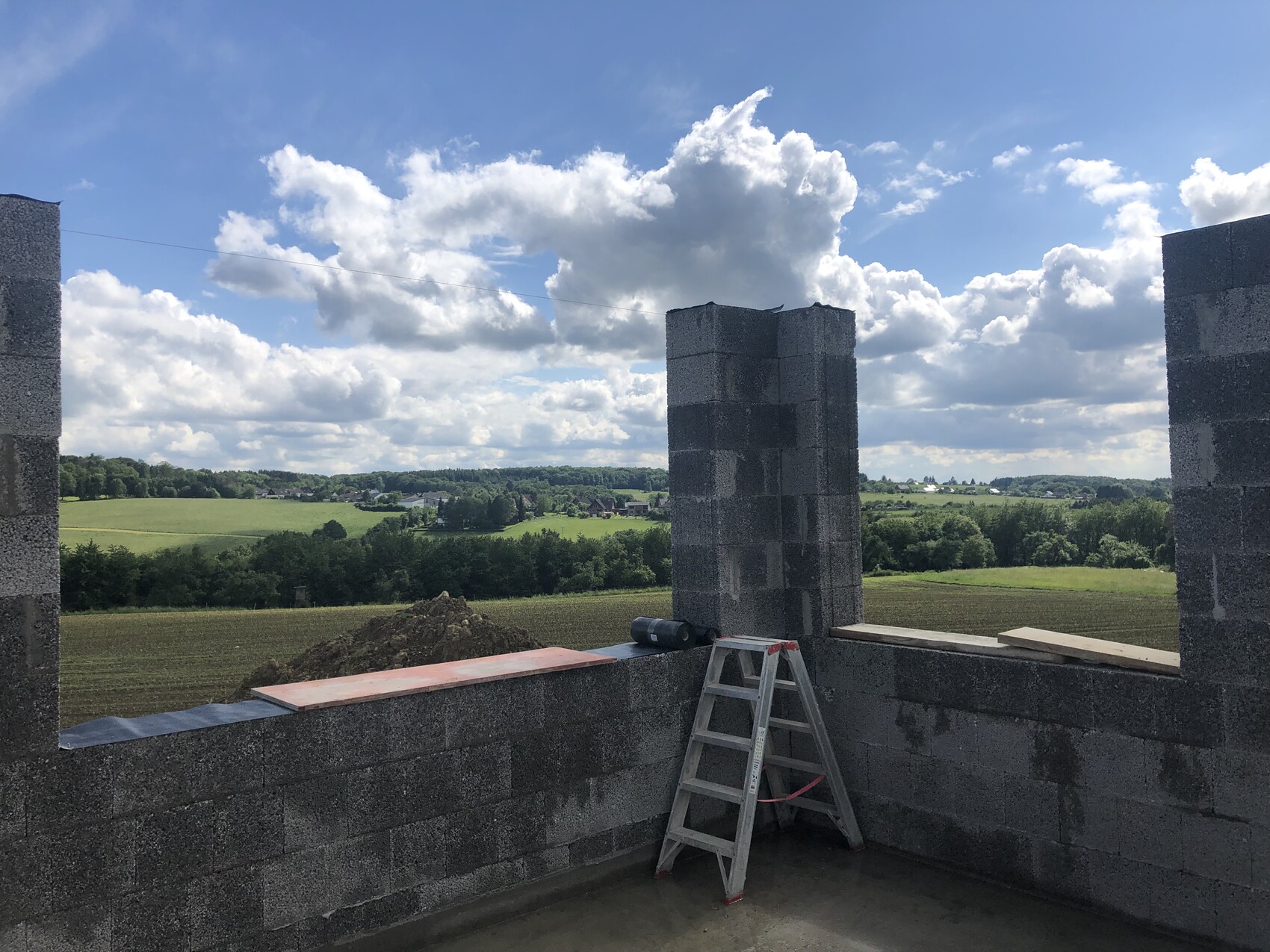 Baustellenansicht: graue Betonblöcke, Leiter, Holzplatten, Blick auf grüne Landschaft.