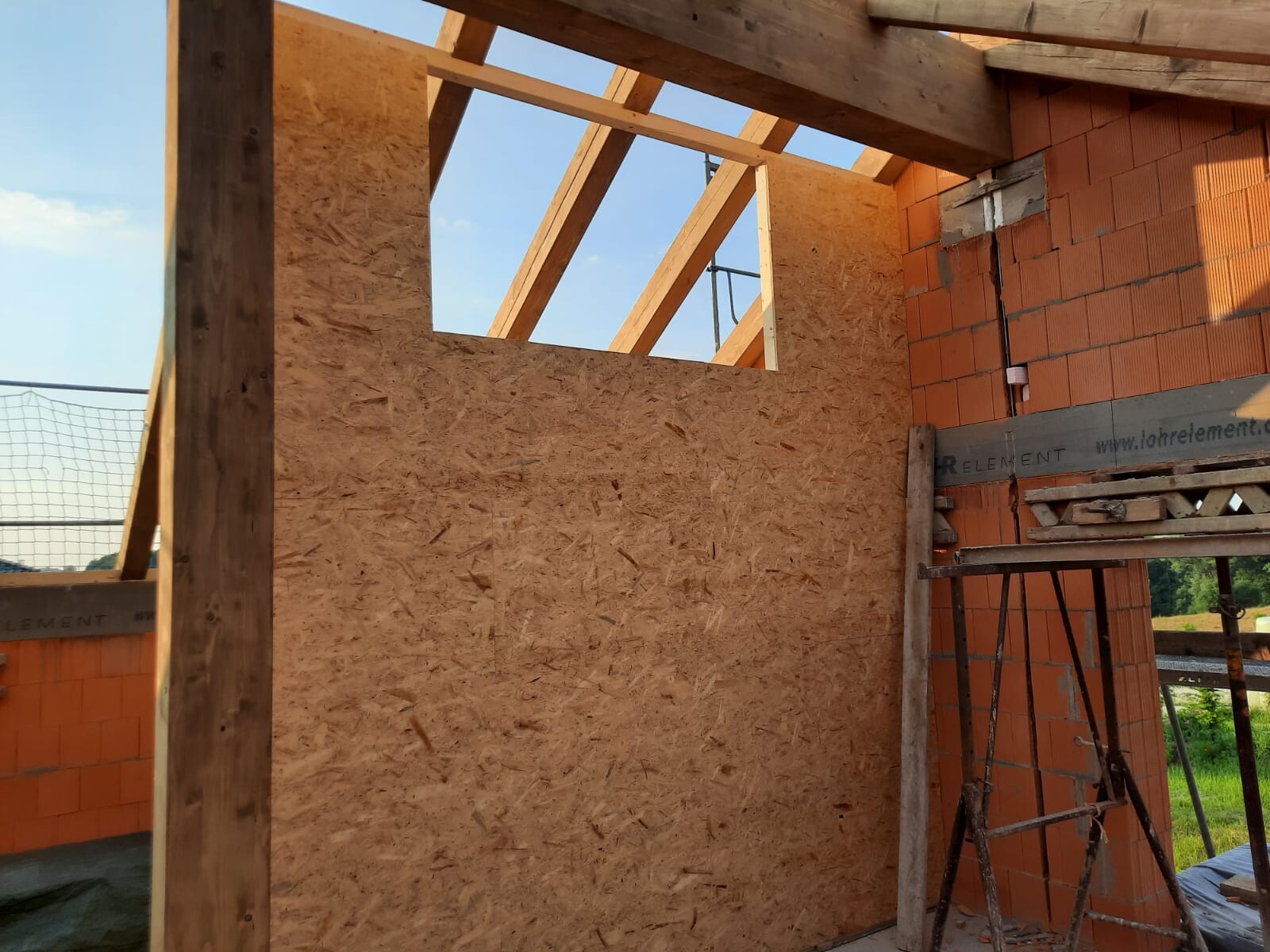 Baustelle mit Holzrahmen und OSB-Wand, orange Ziegelwand und Gerüst, offener Blick nach draußen.