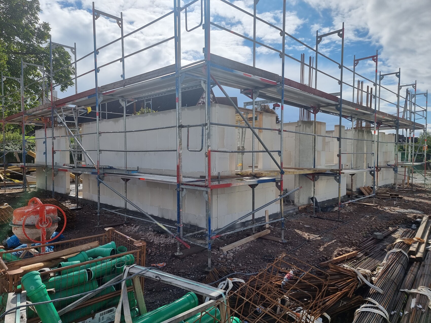 Baustelle mit Gerüst rund um Rohbau aus weißen Betonblöcken; Materialstapel und Betonmischer