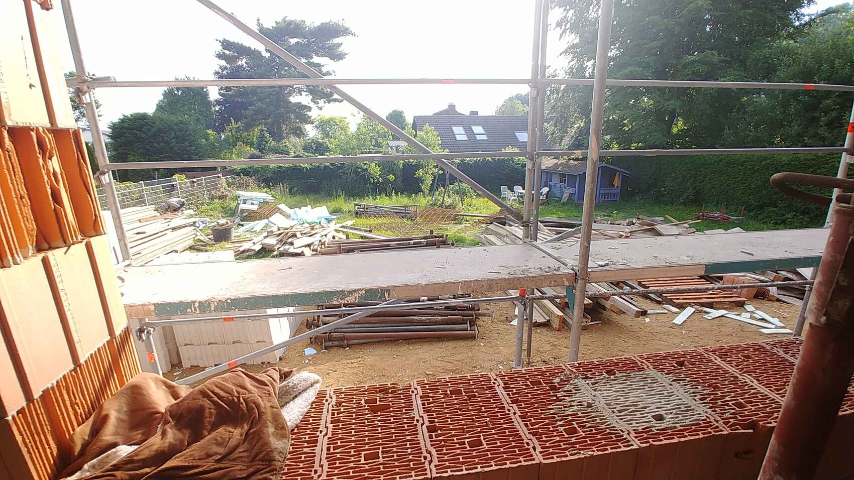Baustelle mit Gerüst und Holzstapeln; Blick von Balkon auf Garten und Haus im Hintergrund