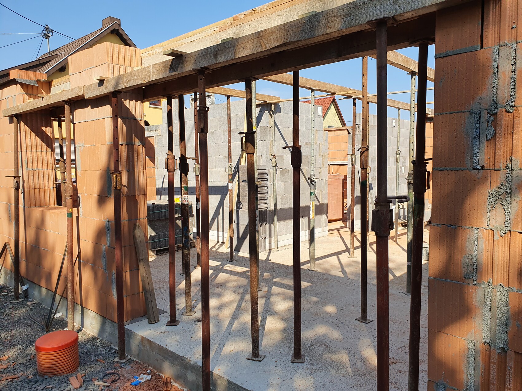 Baustelle mit Stahlstützen, Holzbalken, Ziegelwänden und Betonboden; Baufortschritt sichtbar.