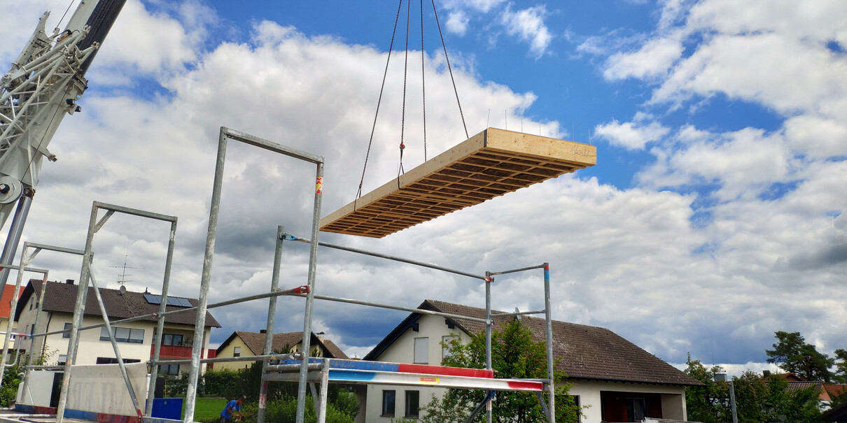 Kran hebt Holzbodenplatte auf Rohbau-Skelett vor Wohnhäusern.