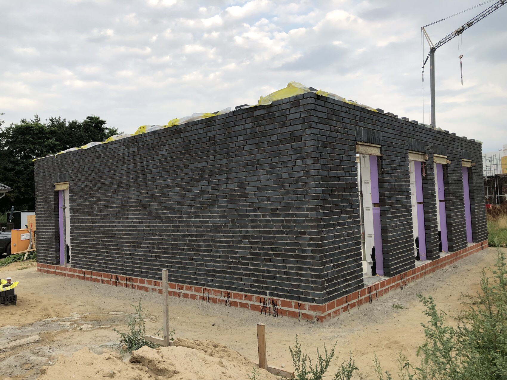 Neubau aus dunklen Ziegeln mit mehreren Türöffnungen und violetten Rahmungen; Kran im Hintergrund.