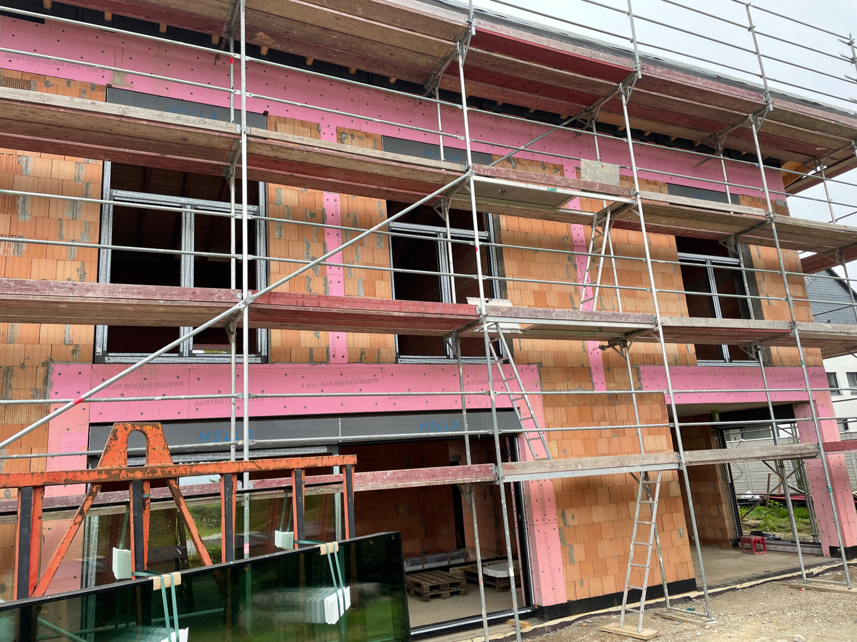 Baustelle: mehrgeschossiges Haus mit Gerüst, pinken Dämmplatten und orange Ziegeln, Leiter.