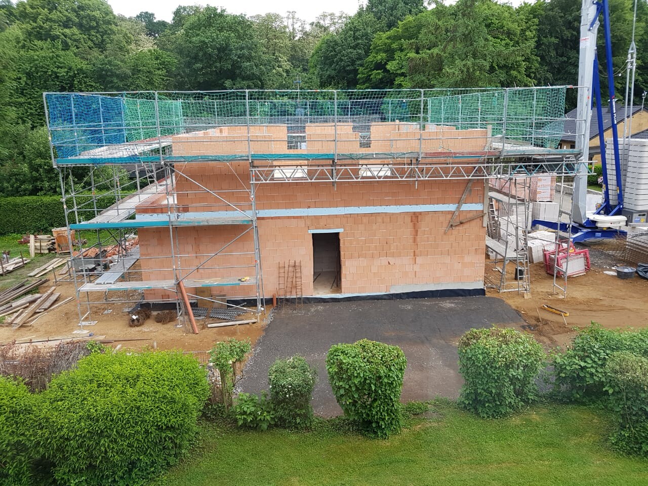 Rohbau aus Ziegeln mit Gerüst umgeben; Eingangstür in der Mitte, Baustellenmaterialien sichtbar.