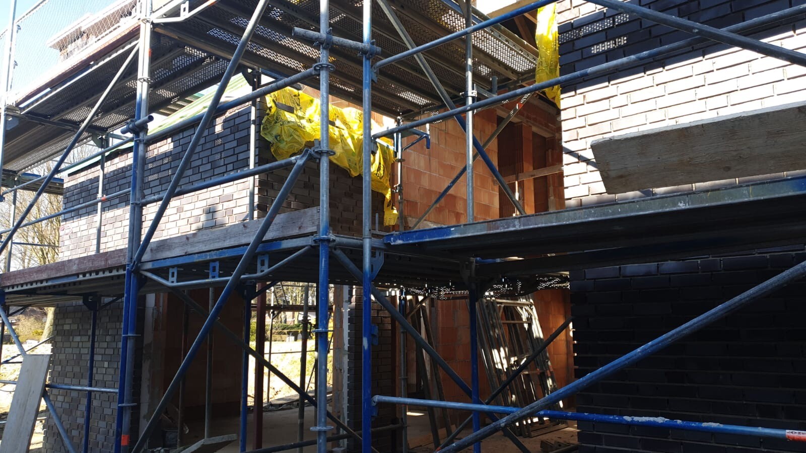 Baustelle: Gerüst um ein gemauertes Gebäude, gelbe Abdeckfolie, Holzbauteile sichtbar.
