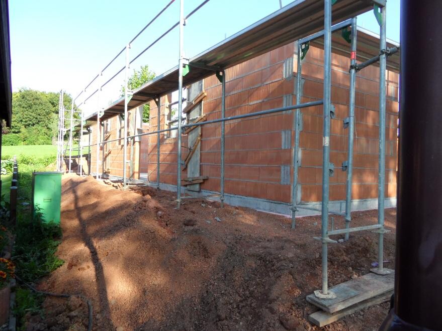 Baustelle mit rotem Ziegelbau und Gerüst, Erdhaufen vor Wand, grüne Landschaft dahinter.