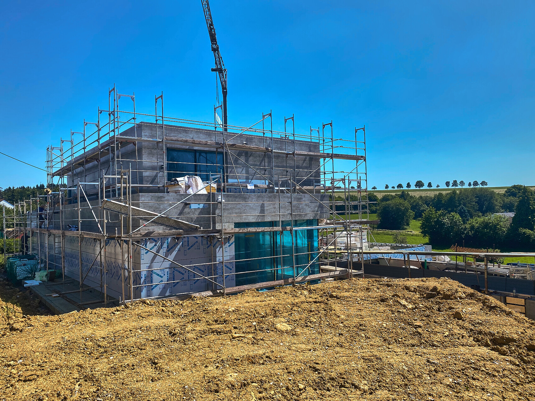 Baustelle mit Gerüst um ein modernes Gebäude, Kran im Hintergrund, Erdhügel im Vordergrund.