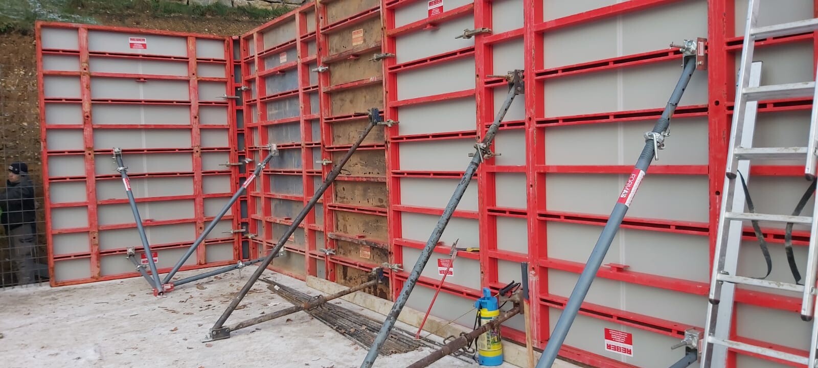 Rote Schalungswände mit Stahlstützen bilden eine Eckwand; Leiter rechts, Bauwerkzeuge am Boden.