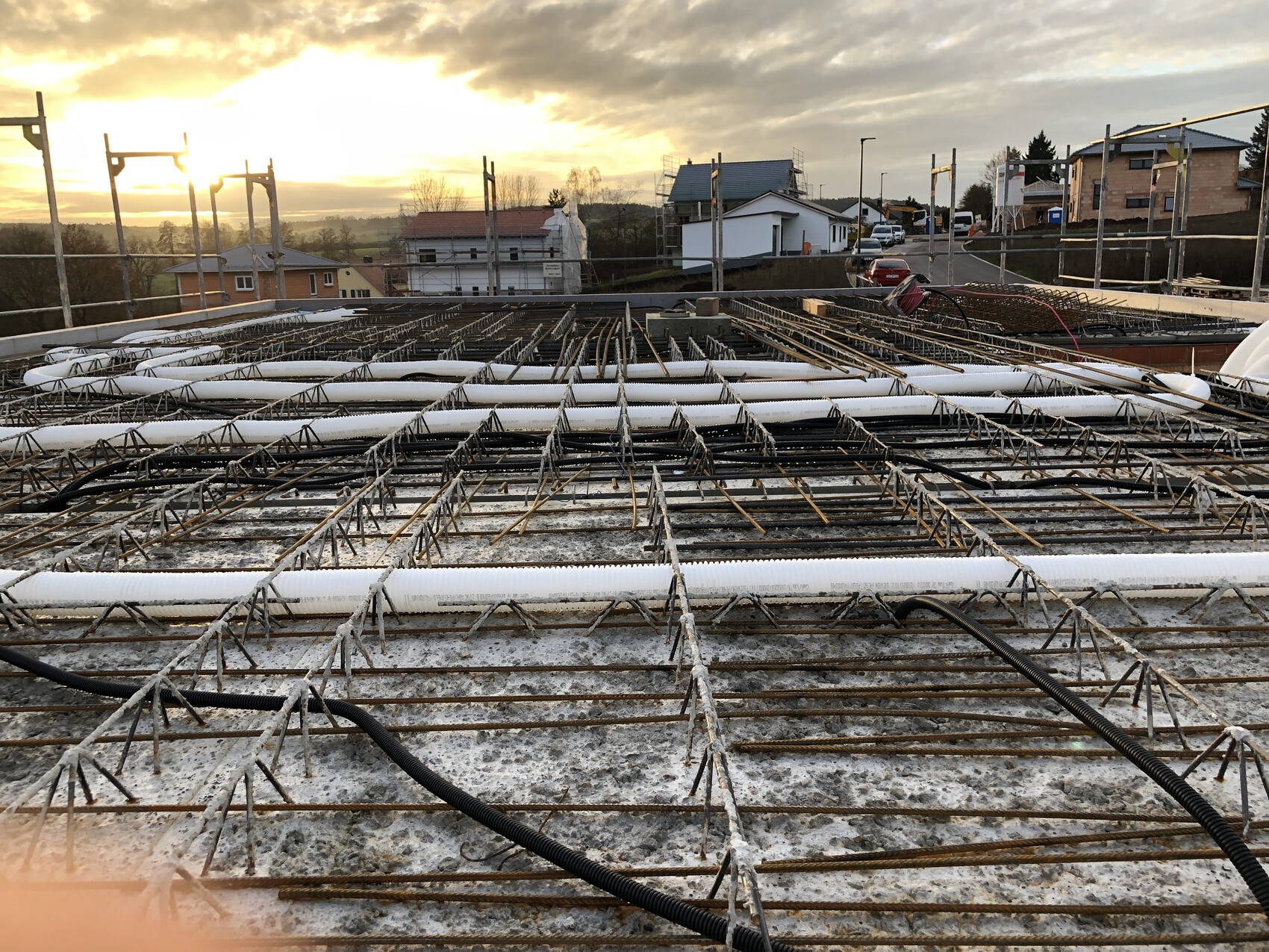 Bewehrungsstahl-Gitter auf Bauplatte; Rohre sichtbar, Häuser bei Sonnenuntergang im Hintergrund