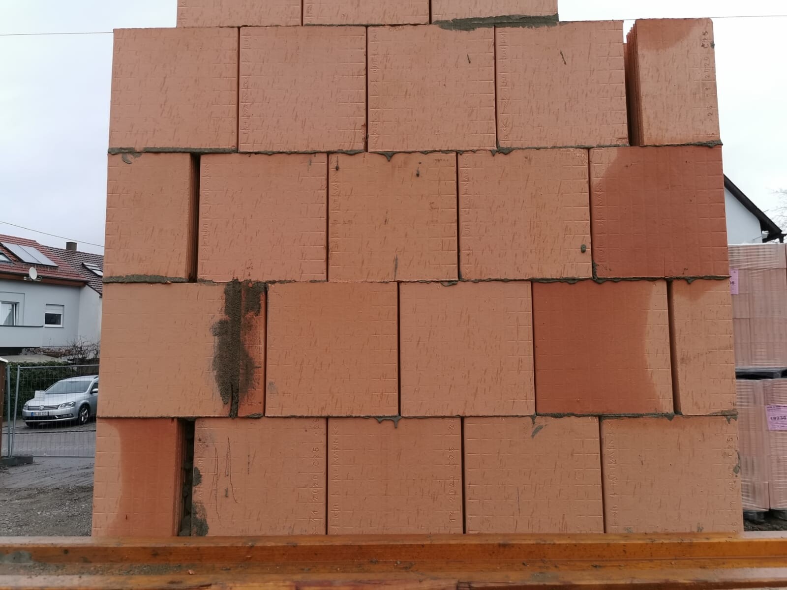 Große orange Ziegelsteine stapeln sich zu einer Wand auf einer Baustelle; Häuser im Hintergrund.