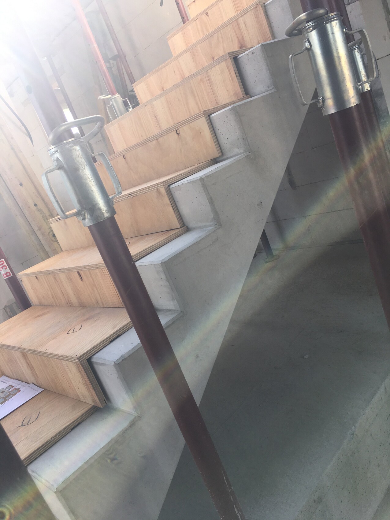 Baustelle: Holzplatten stapeln sich neben Betontreppe; zwei Metallrohre mit Clips stehen davor.