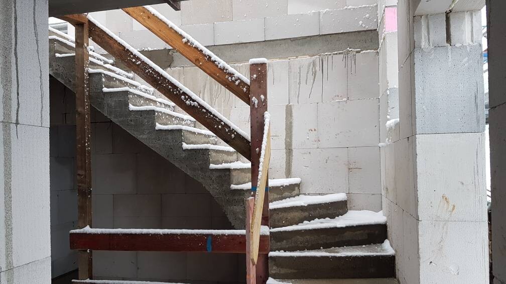 Unfertige Baustelle mit kurviger Betontreppe, Holzgeländer und Schnee auf den Stufen.
