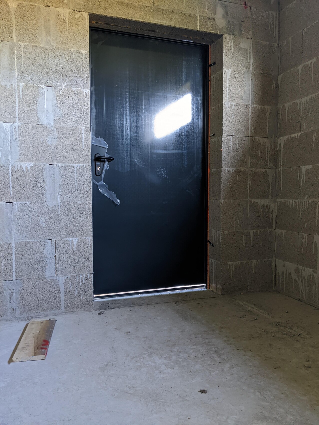 Dunkle Metalltür im Rohbau, Betonwände, beschädigte Griffabdeckung, staubiger Boden