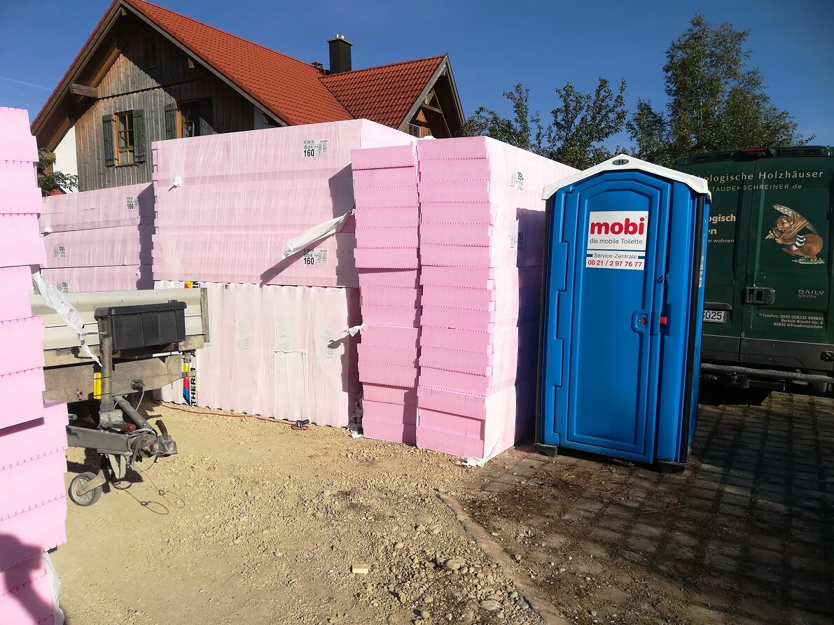 Baustelle mit rosa Dämmplatten, blaues Porta-Potty und Baufahrzeuge