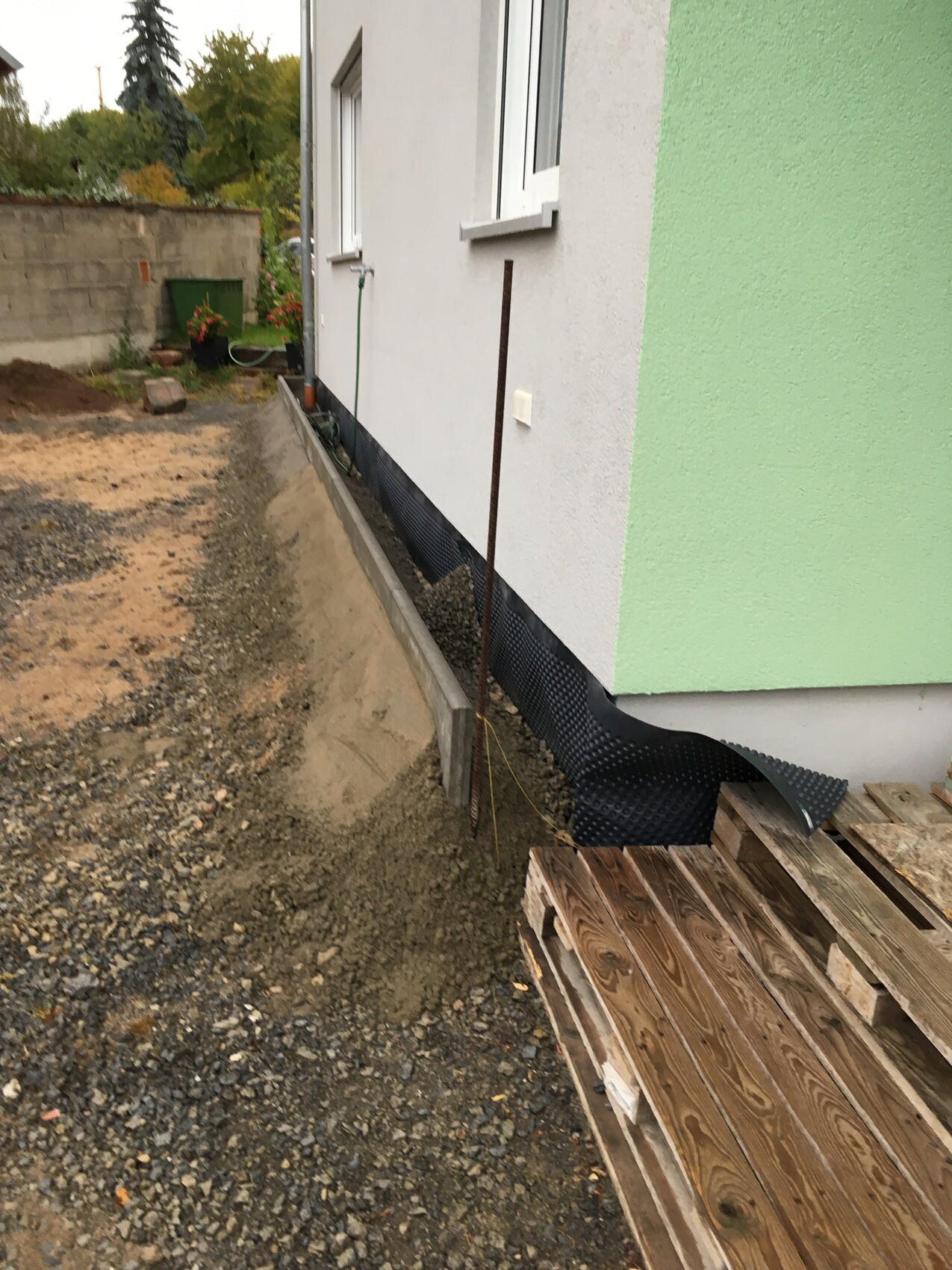 Baustelle am Haus: Graben am Fundament, schwarze Abdichtbahn, Holzpaletten neben der Wand.
