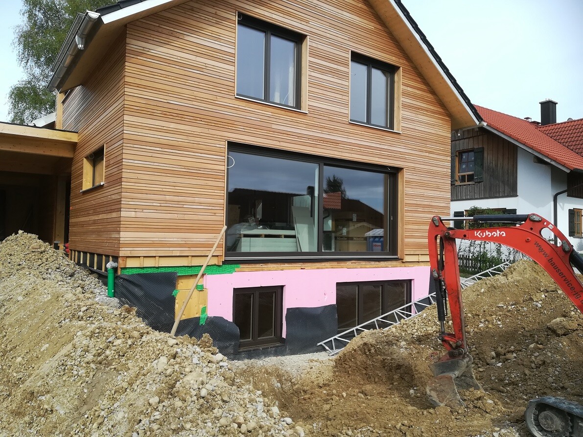 Mehrstöckiges Holzhaus im Bau mit großen Fenstern, Erdarbeiten und rotem Bagger im Vordergrund.
