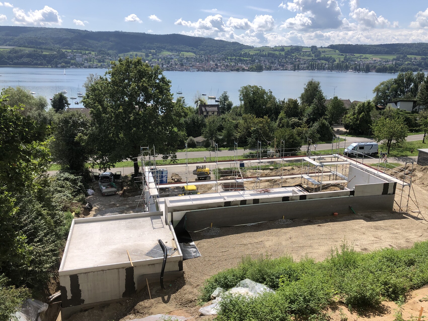 Baustelle mit Betonfundamenten und Gerüst, am Seeufer mit Booten und Bergen im Hintergrund.