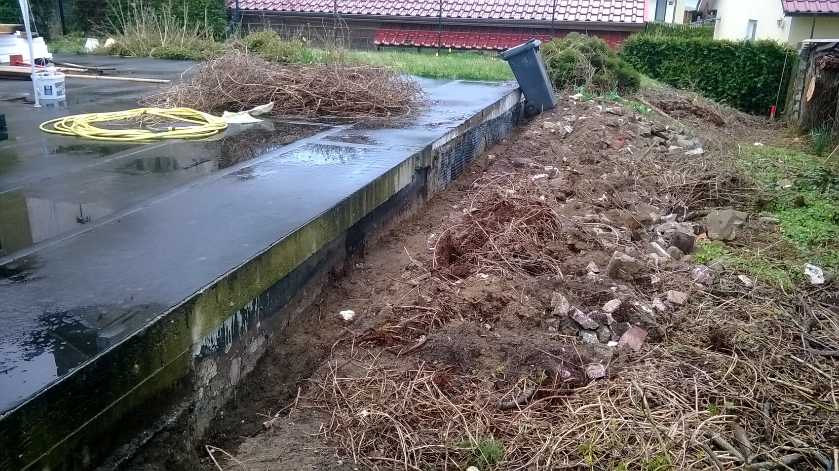 Nasse Betonplatte mit gelbem Schlauch, Astwerk und Schutt neben Müllcontainer.