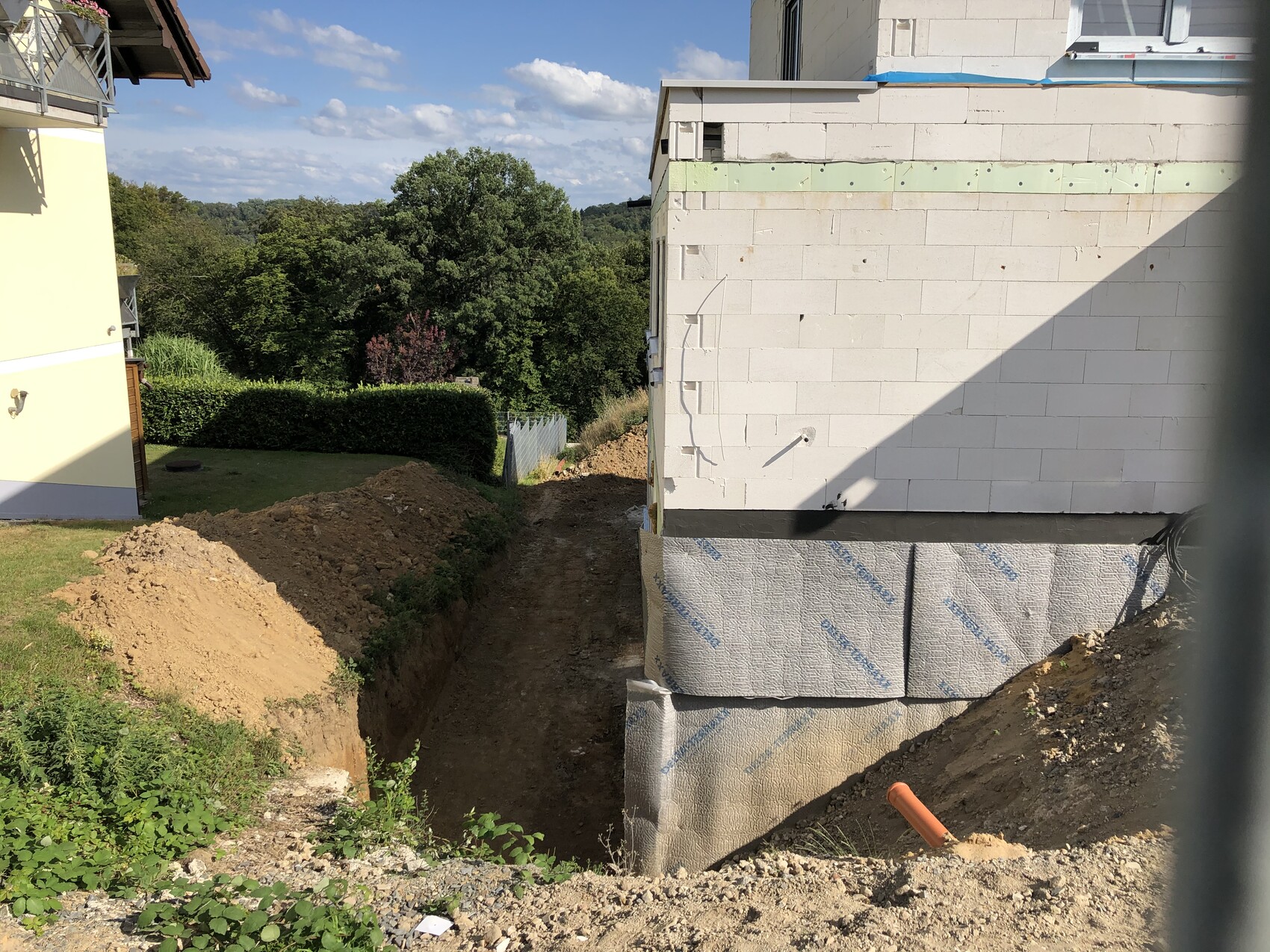 Baustelle neben Haus: Graben am Fundament, weiße Blöcke, Dämmplatten, orange Rohr vorn.