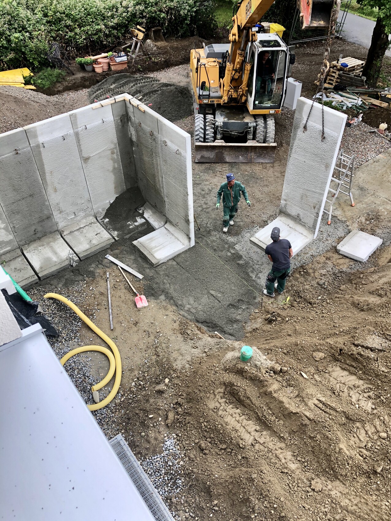 Blick von oben auf eine Baugrube mit Betonwänden, Bagger, zwei Arbeiter in Arbeitskleidung