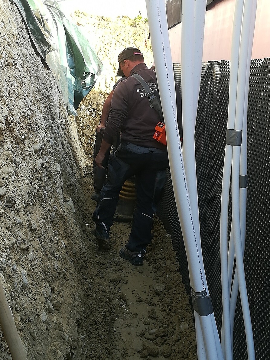 Bauarbeiter in dunkler Arbeitskleidung kniet im engen Graben neben Rohbauwand und Rohren
