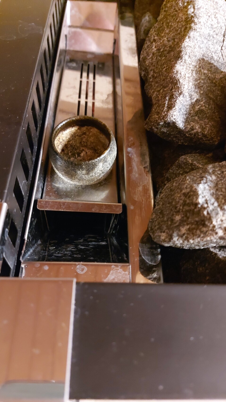 Runder Metall-Aschebehälter mit Asche, auf brauner Ablage neben Steinblock eines Kamins.