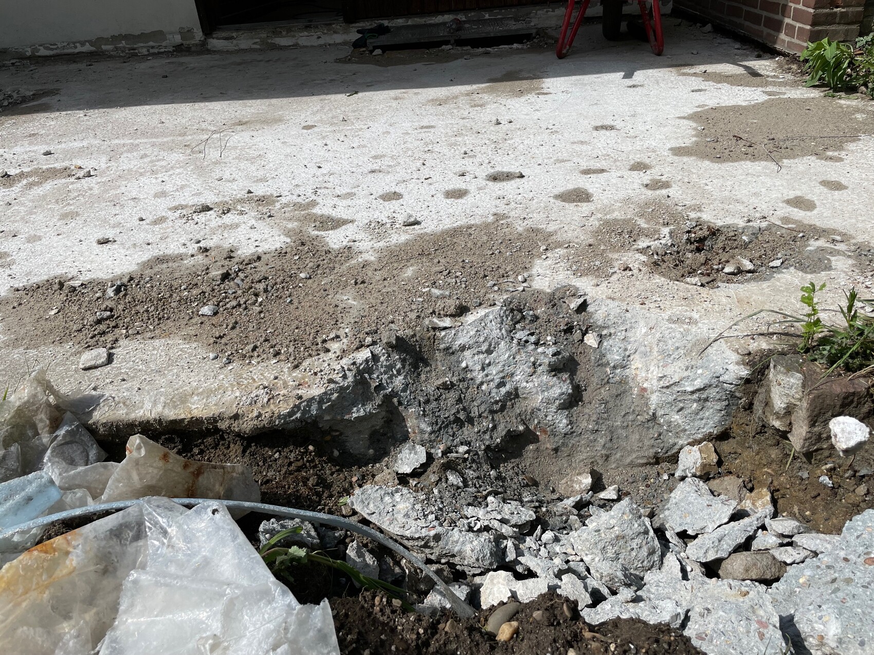 Aufgebrochene Betonfläche mit Graben, Kies und Geröll; links Müll, rechts Pflanzen.