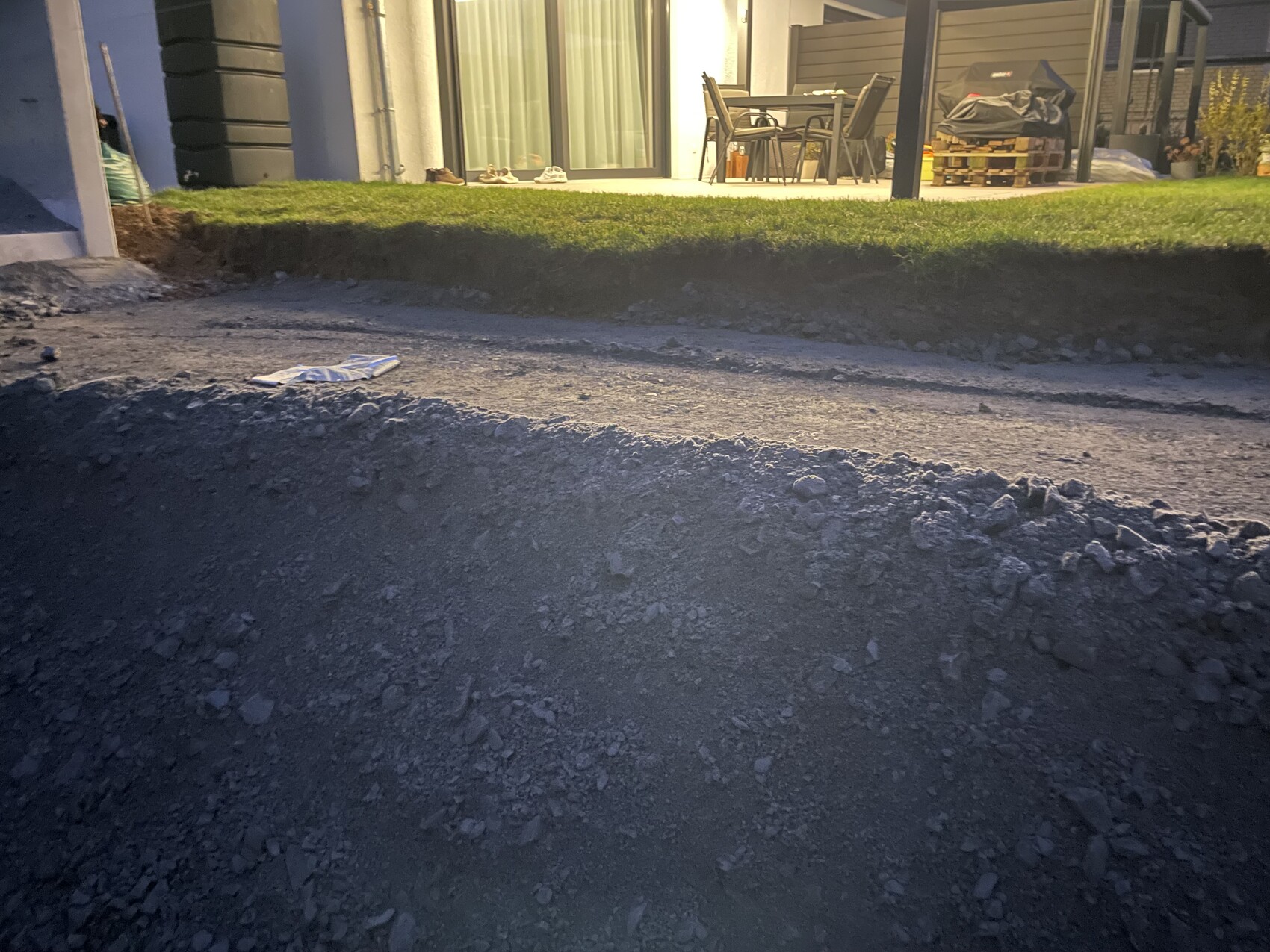 Baugrube vor dem Haus, Erde und Kies, Terrasse mit Gartenmöbeln im Hintergrund.