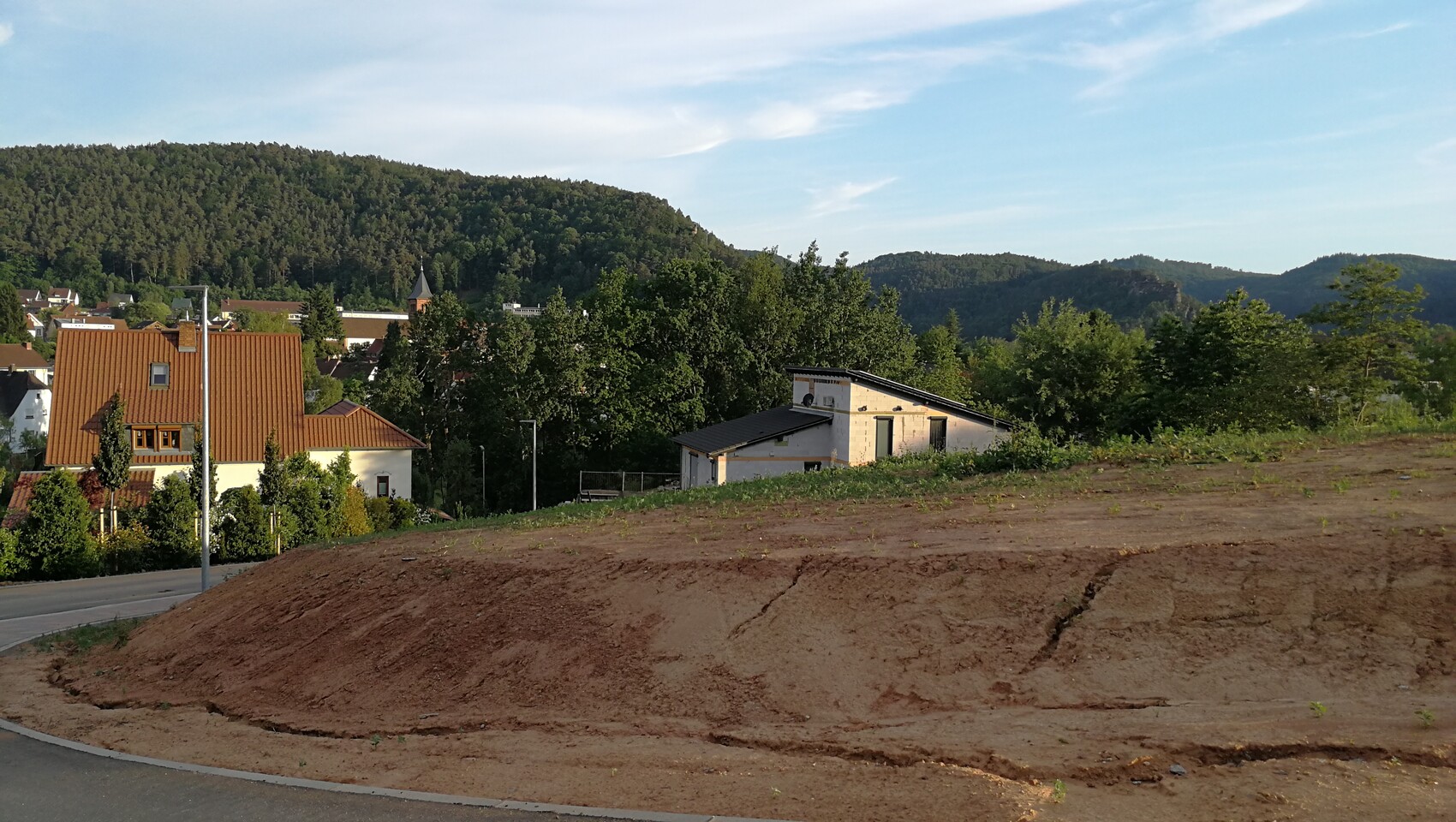 Bauanlage und halbfertiges Haus im Vordergrund, links rote Dächer, Waldhügel im Hintergrund.