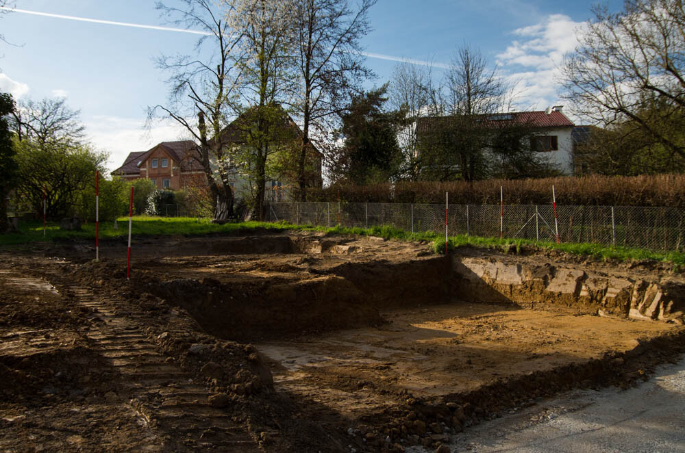 Ausgehobenes Baugrundstück mit Grube, Zaun und Häusern im Hintergrund