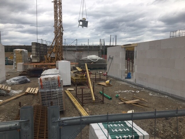 Kran hebt Last auf Baustelle; unfertige Betonwände und Baumaterialien liegen herum.