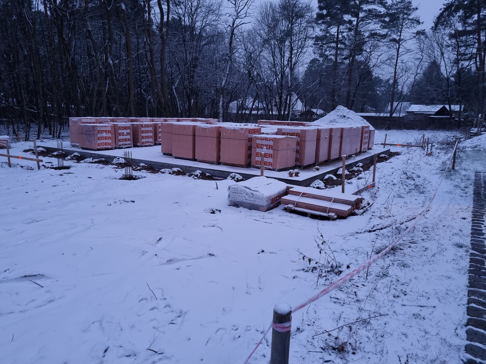 Winterbaustelle mit verpacktem Baumaterial auf Betonplatte; Schnee und Wald im Hintergrund.