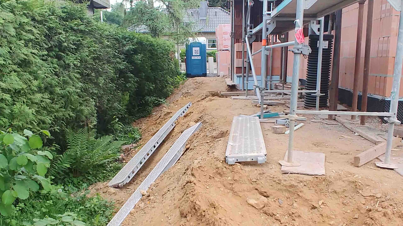 Baustelle mit Graben, Baugerüst, ToiToi-WC, Metallplatten und Erdreich neben grünem Gebüsch.
