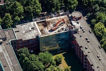 Luftaufnahme eines Gebäudes mit Dachbaustelle und Baumaschine auf dem Dach, umliegende Bäume.