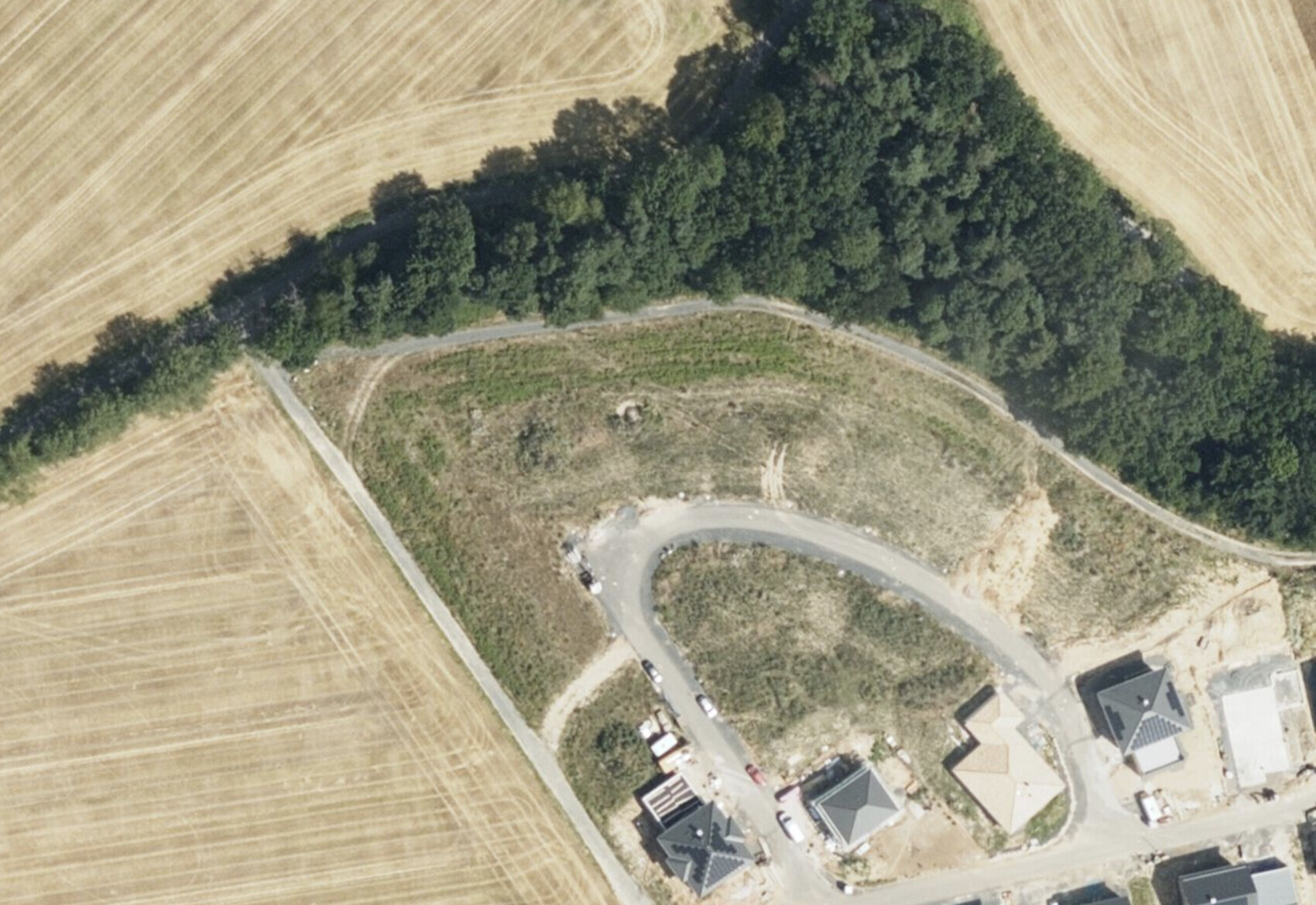 Kurvenreiche Straße um ein grasiges Plateau, umgeben von Feldern und Wald; Häuser unten rechts.