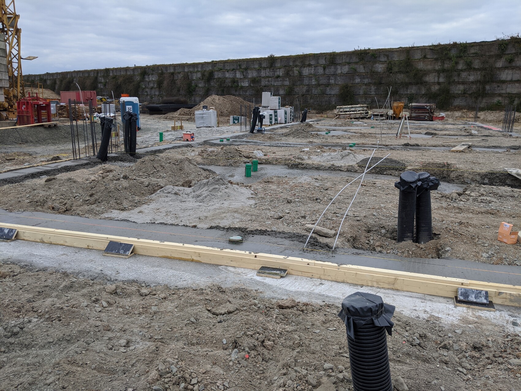 Baustelle mit Gräben, schwarzen Rohren, Holzformen und Baumaterialien, Mauer im Hintergrund.