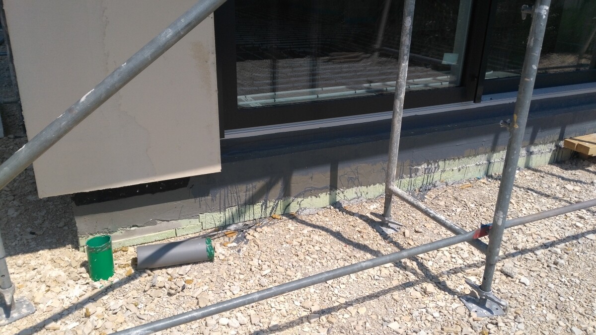 Baustelle am Gebäude: Gerüstrohre, Kiesboden, grüne Farbdose neben Fenster.
